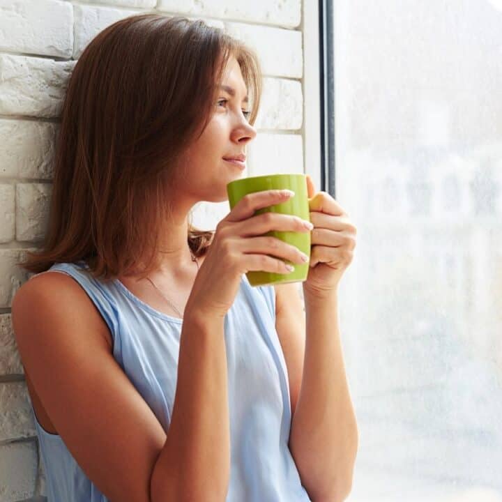 A woman in a light blue sleeveless top sits by a window, holding a green mug and looking outside thoughtfully.
