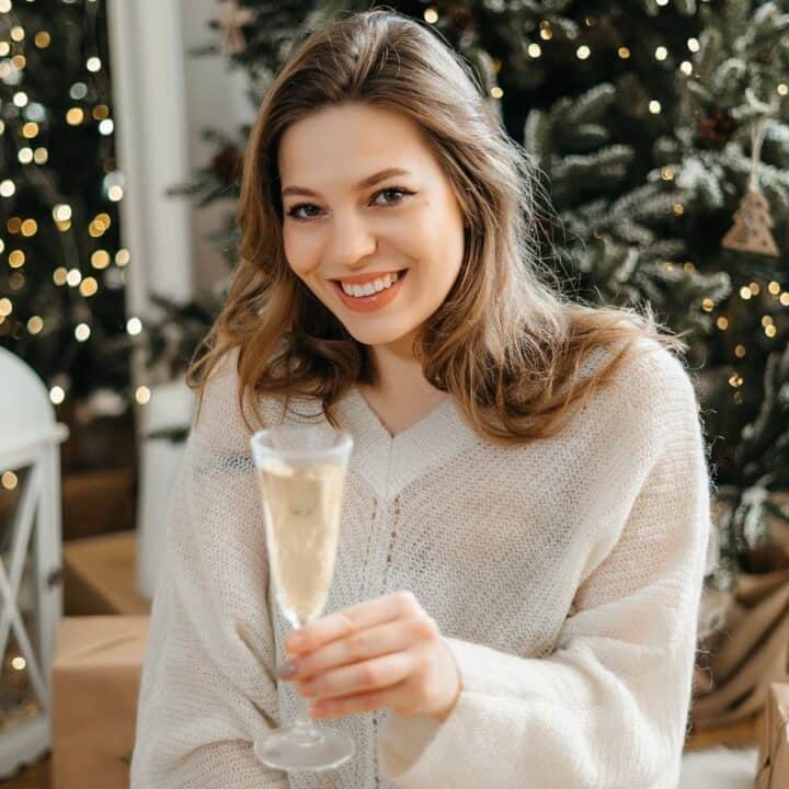 A woman in a white sweater holds a glass of champagne and smiles, sitting in front of decorated Christmas trees and wrapped gifts.