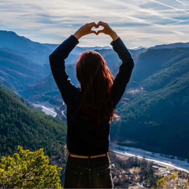 A person stands on a mountain overlook forming a heart shape with their hands, facing a scenic landscape of forests, rivers, and distant mountains under a blue sky.