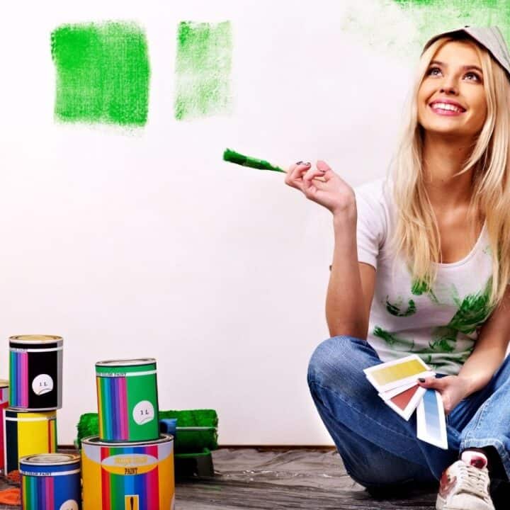 A woman sits on the floor with paint cans and swatches, holding a paint roller with green paint. Green paint is partially applied to the wall behind her.
