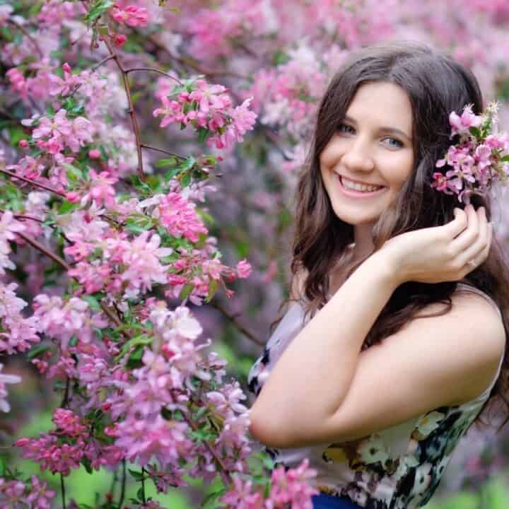 A woman with long brown hair stands among blooming pink flowers, smiling and holding a small branch of blossoms near her face.