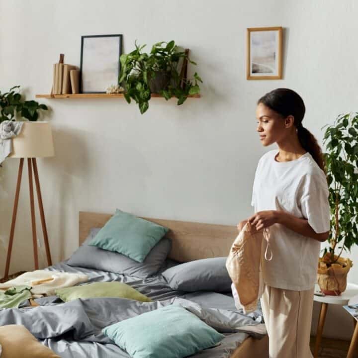 A woman stands beside an unmade bed in a bright bedroom with several potted plants, scattered clothes, books, and framed pictures on the wall.