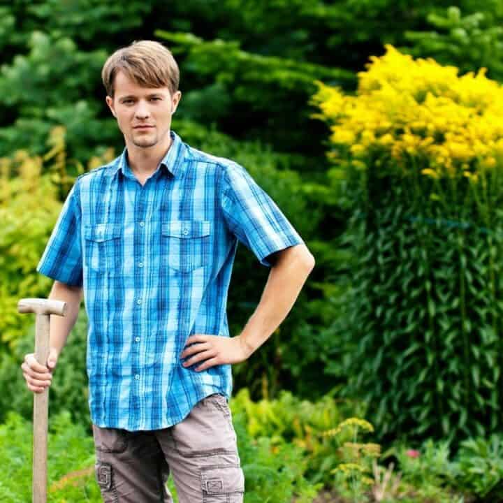 A man in a blue plaid shirt stands in a garden holding a spade, surrounded by green plants and blooming yellow and pink flowers.