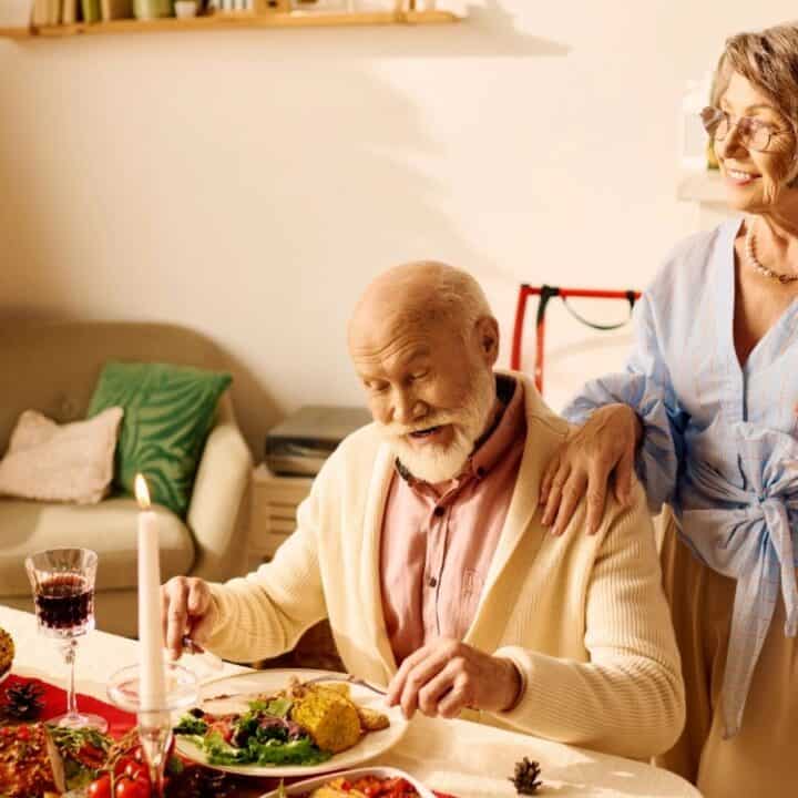An elderly man sits at a festive dinner table while an elderly woman stands beside him holding a glass of red wine. A decorated Christmas tree is visible in the background.