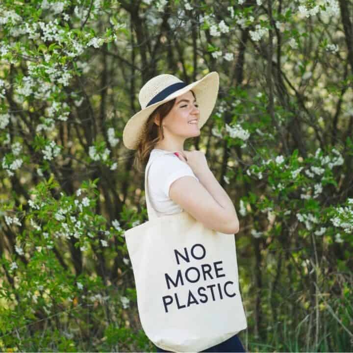 Woman standing outdoors among green trees, wearing a hat and holding a tote bag with the words "NO MORE PLASTIC" printed on it.