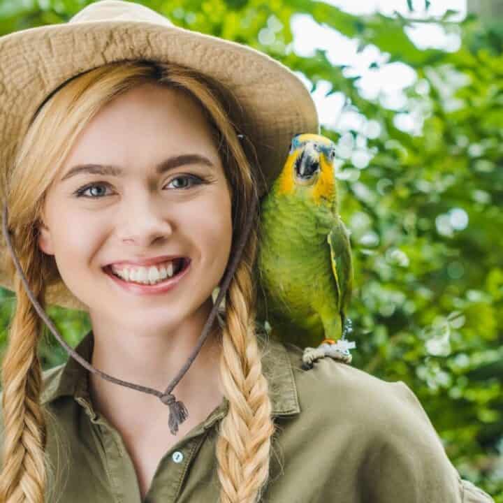 Smiling woman with blonde braids wearing a hat and green shirt standing outdoors with a green parrot perched on her shoulder.