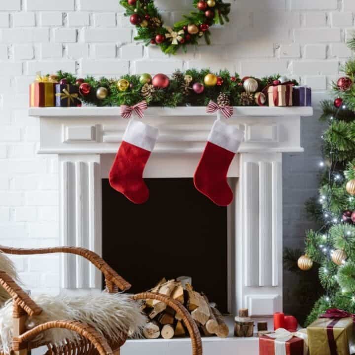 A woman decorates a Christmas tree next to a fireplace with stockings, presents, a wreath, and festive ornaments in a cozy living room.