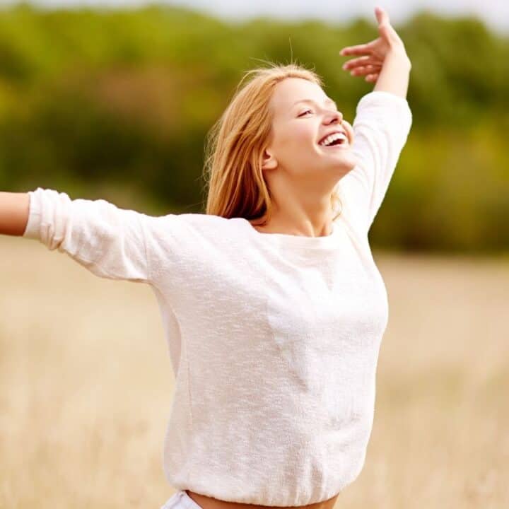 A person standing in a field with arms outstretched, smiling and looking upward, with trees and sky in the background.