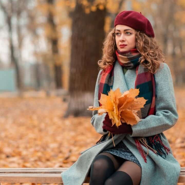 A woman wearing a coat, scarf, and beret sits on a bench in a park holding yellow autumn leaves. Fallen leaves cover the ground and trees with bare branches are in the background.