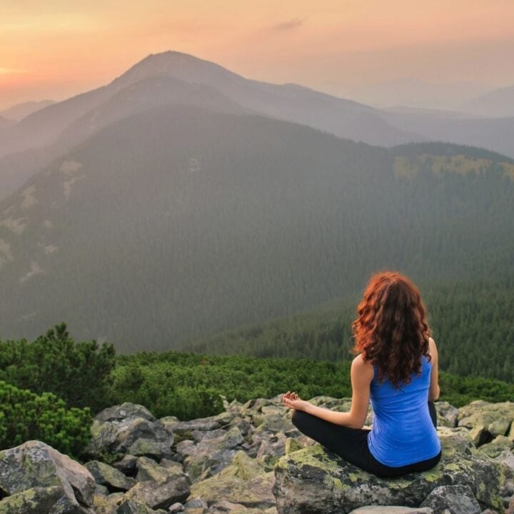 A person sits on rocky ground facing mountains and a sunset, meditating or relaxing, surrounded by green trees and hills.