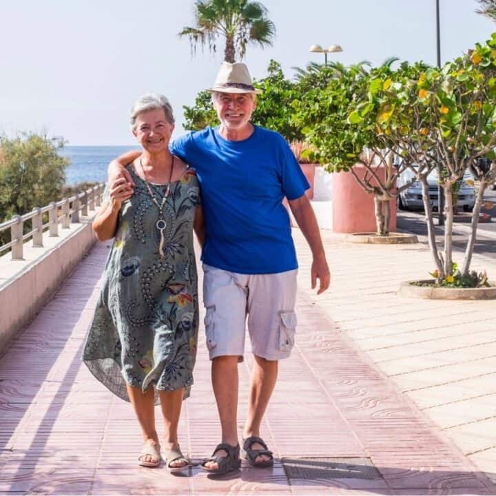 Two older adults walk arm in arm on a sunny promenade near the ocean, surrounded by palm trees and parked cars.