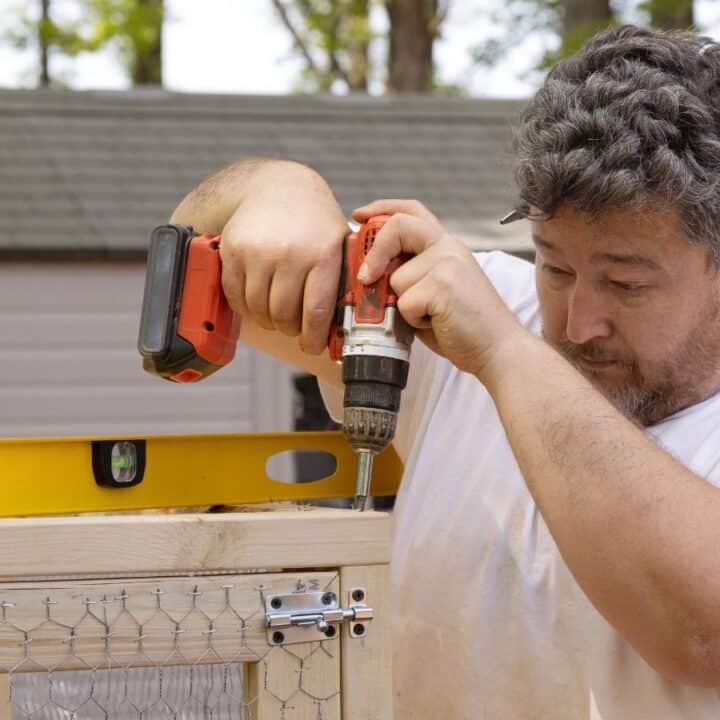 A man uses a power drill to secure a hinge to a wooden frame outdoors, with a yellow level resting on top of the structure.