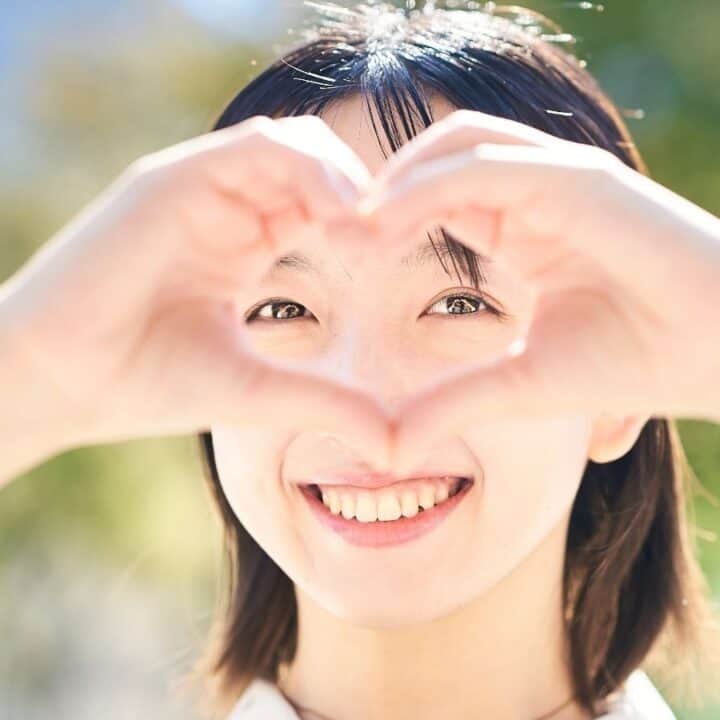 A person smiles while holding their hands in the shape of a heart in front of their face, outdoors with a blurred background.