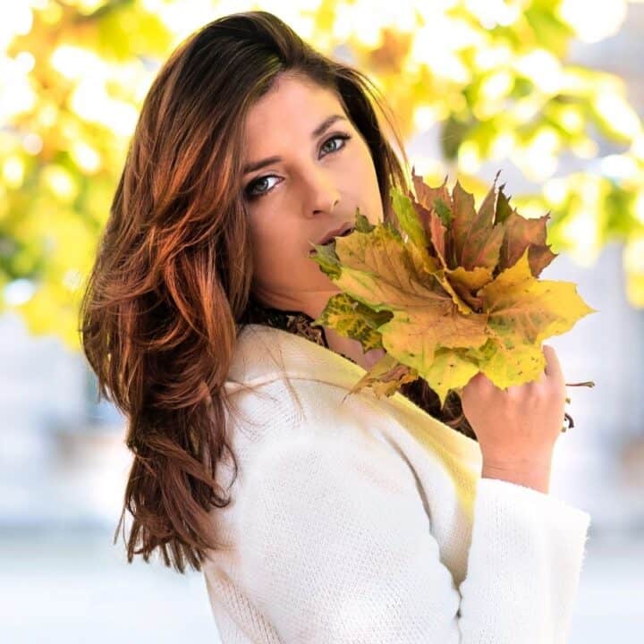 A woman with long brown hair holds a bunch of autumn leaves near her face while standing outdoors, with sunlight and blurred greenery in the background.