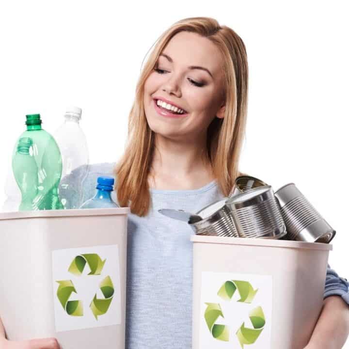 A woman holds two recycling bins, one filled with plastic bottles and the other with metal cans, standing against a white background.
