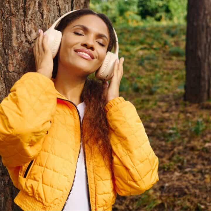 A woman in a yellow jacket listens to headphones and smiles while standing next to a tree in a wooded outdoor area.