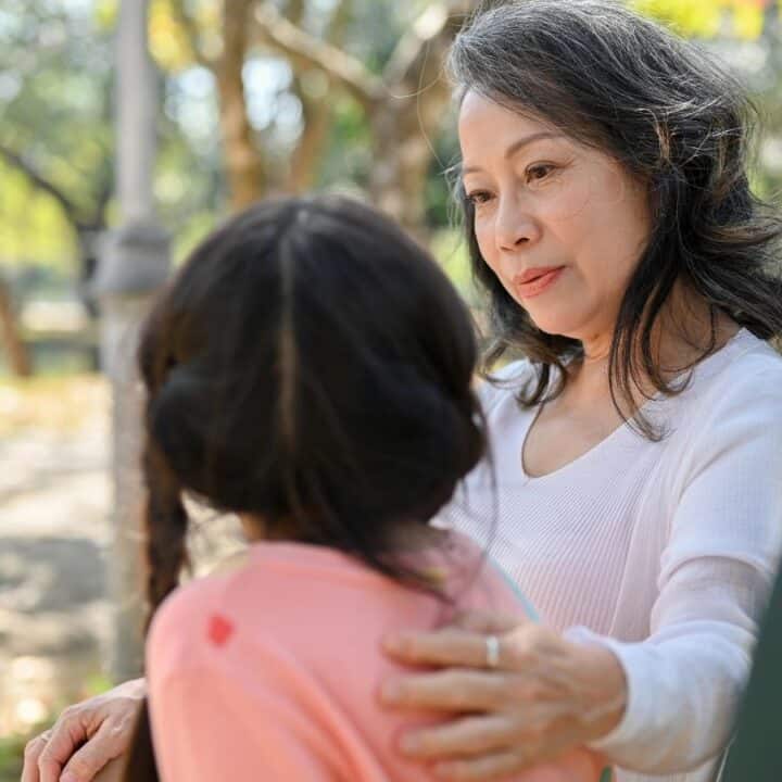 An older woman sits outdoors, gently holding and talking to a young girl with braided hair, surrounded by trees and sunlight.