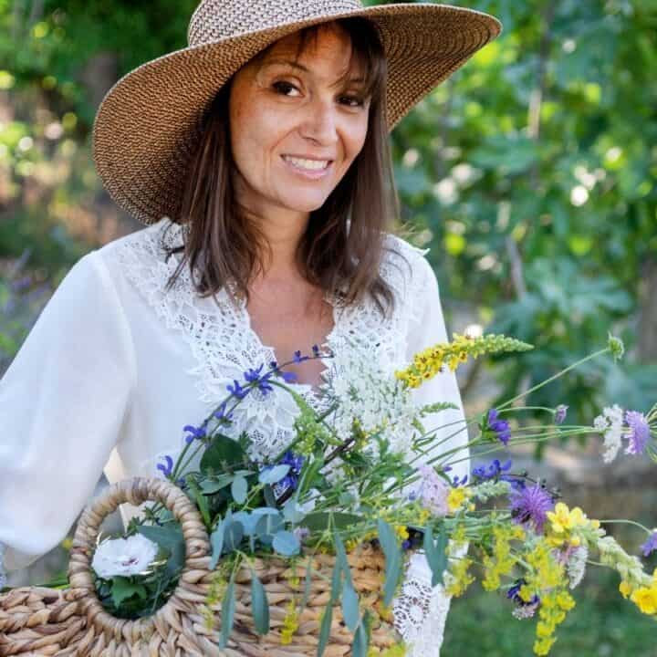 Woman in a straw hat and white blouse stands outdoors, holding a woven basket filled with assorted wildflowers. Greenery and purple flowers are visible in the background.