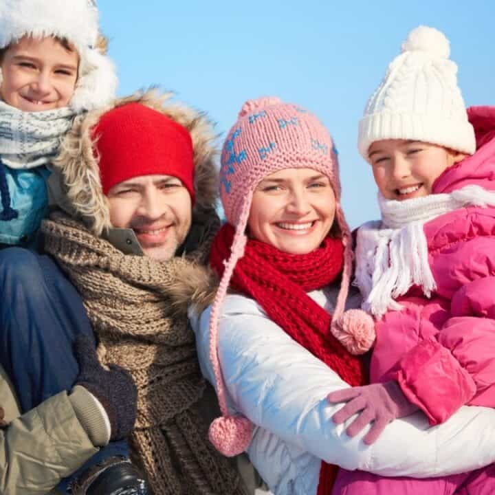 A family of four dressed in winter clothing, smiling outdoors on a snowy day. The parents are holding their two children.