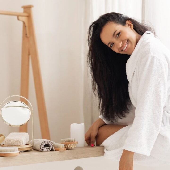 A woman in a white bathrobe sits on the edge of a bathtub, smiling, with various bath and beauty products arranged on the tub's ledge.