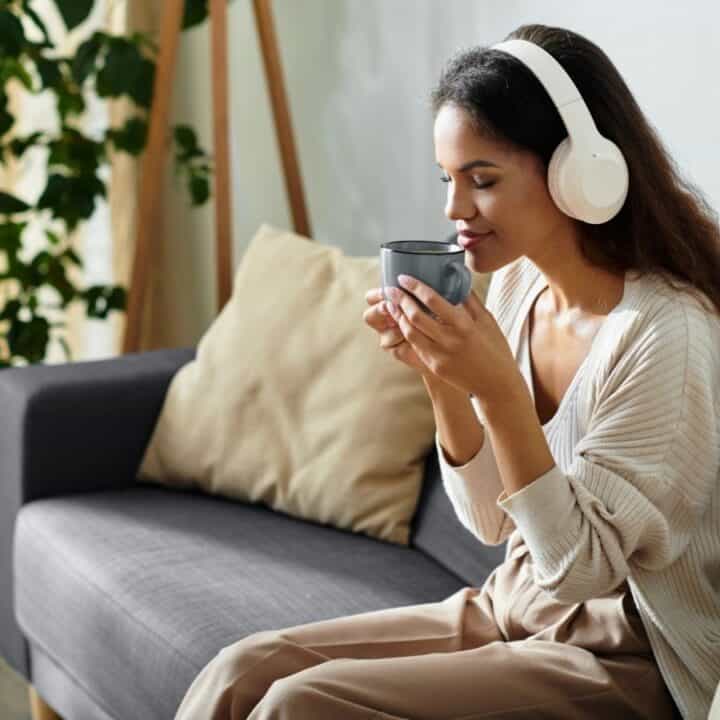 Woman sitting on a sofa, wearing headphones, and holding a mug with both hands, appearing relaxed. There are plants and cushions in the background.