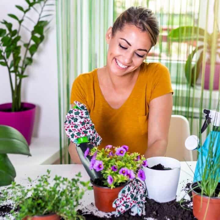 A woman in a yellow shirt smiles while potting flowers at a table with gardening supplies and potted plants around her.