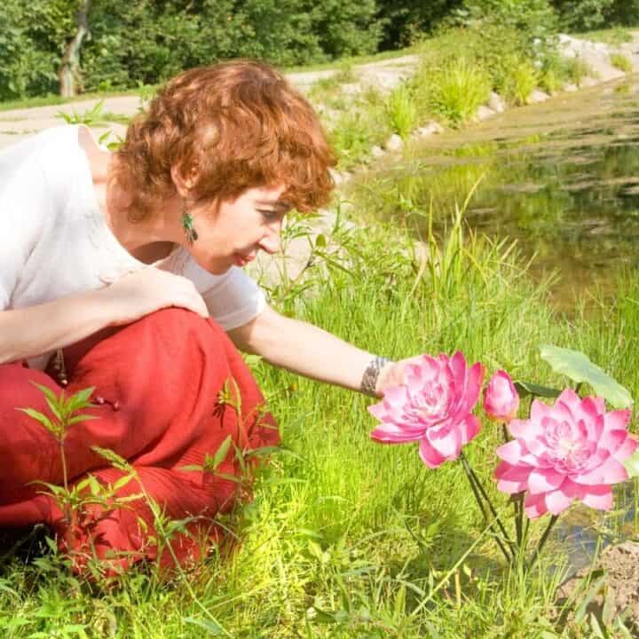 A woman with short red hair and red pants kneels by a pond, reaching out to touch two large pink flowers growing near the water's edge.