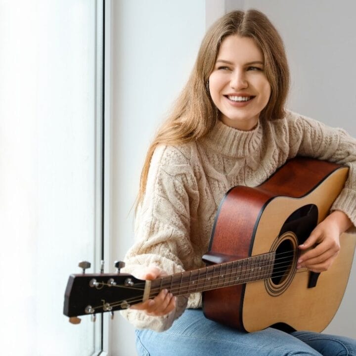 A woman with long hair sits by a window, smiling and playing an acoustic guitar while wearing a beige sweater and jeans.
