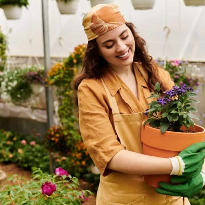 A woman wearing a headscarf, gloves, and apron smiles while holding a potted flowering plant in a greenhouse filled with various hanging and potted plants.