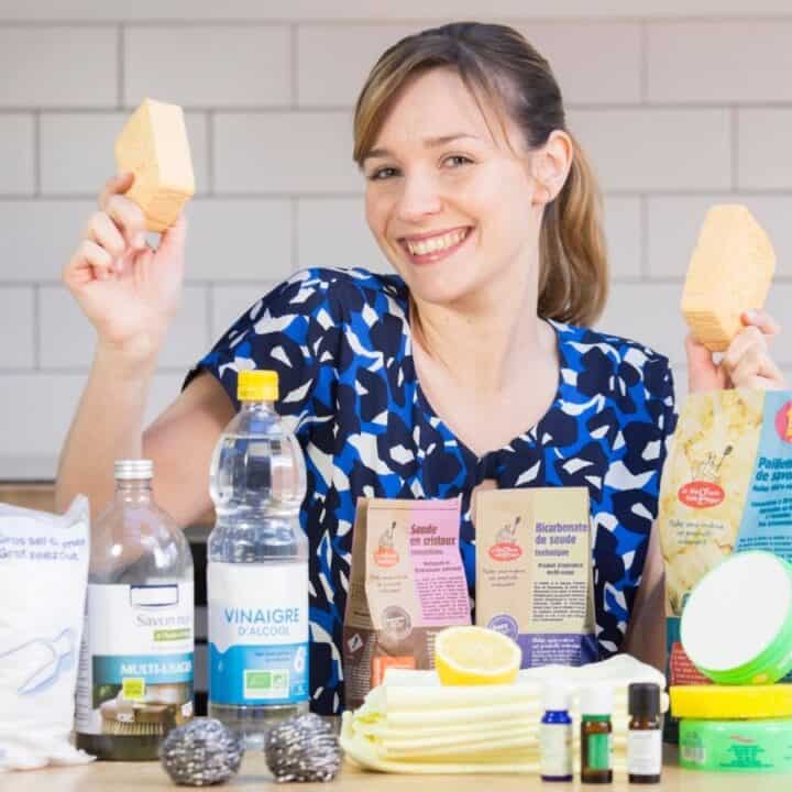 A woman in a kitchen holds up two sponges and smiles. In front of her are various cleaning supplies, including vinegar, baking soda, soaps, brushes, cloths, and essential oils.