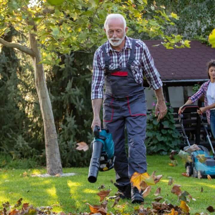 An older man uses a leaf blower on a lawn while a younger woman mows the grass in the background on a sunny day.