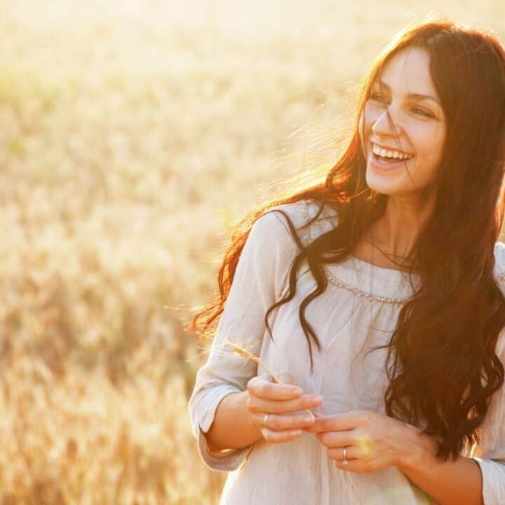 A woman with long brown hair smiles and stands in a sunlit field, wearing a light-colored long-sleeve blouse, holding a stem of grass.