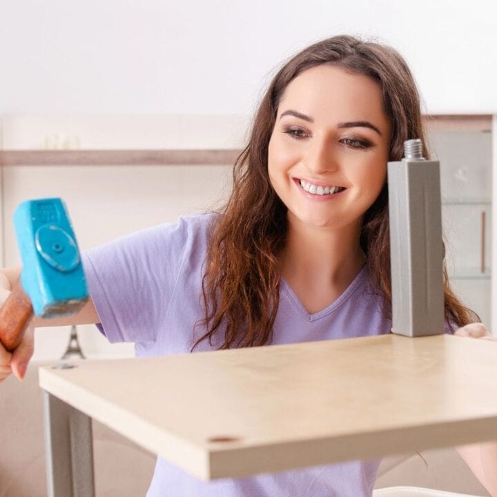 Woman assembling furniture, holding a blue hammer and attaching a metal leg to a wooden tabletop in a bright room.