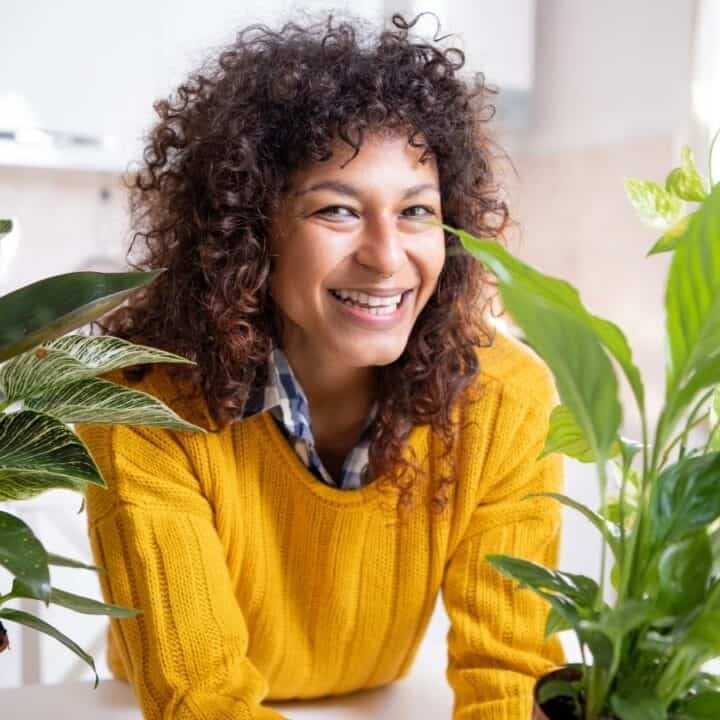 A person with curly hair in a yellow sweater smiles while sitting at a table with two potted green houseplants in a bright kitchen.