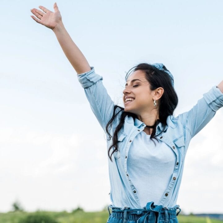 A woman outdoors raises her arms and smiles, wearing a light blue shirt and denim jacket, with a blue sky and green landscape in the background.