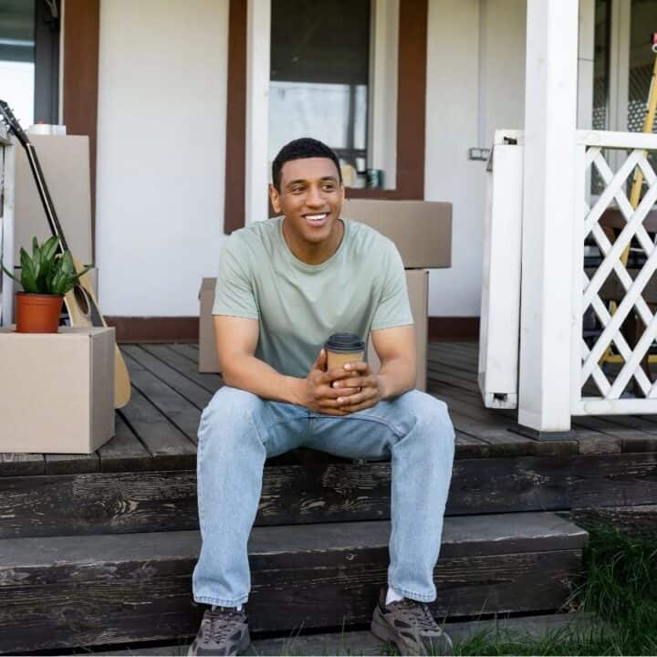 A man sits on the steps of a porch holding a coffee cup, surrounded by moving boxes and a plant, with a house and some outdoor furniture in the background.