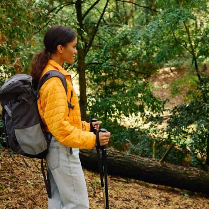 A person wearing a yellow jacket and backpack stands with trekking poles in a forest surrounded by green trees.