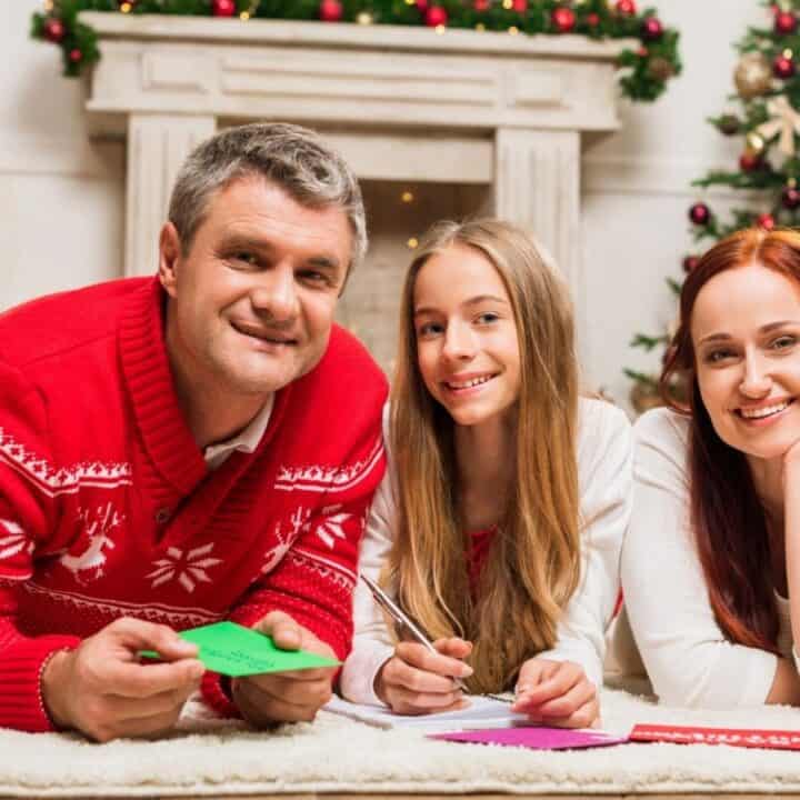 A man, a girl, and a woman lie on the floor in front of a decorated Christmas tree and fireplace, smiling and holding colored paper.