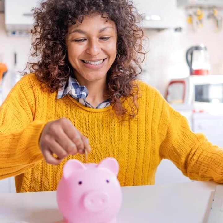 A person in a yellow sweater puts a coin into a pink piggy bank while sitting at a table with papers and a laptop in a bright kitchen.