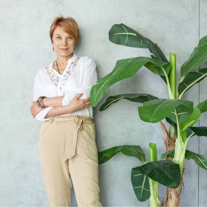 A woman in a white blouse and beige pants stands with arms crossed next to a large green potted plant against a light gray wall.