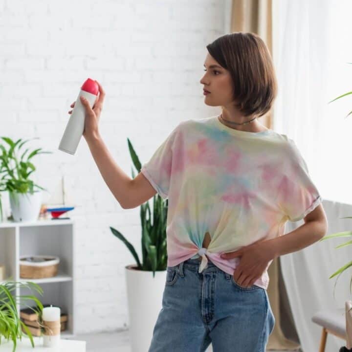 A woman holding an air freshener spray stands in a living room surrounded by various potted plants, looking at the spray can in her hand.
