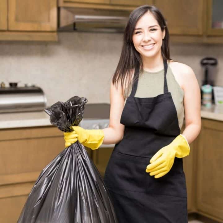 A woman wearing yellow gloves and a black apron holds a large black garbage bag in a kitchen, smiling at the camera.
