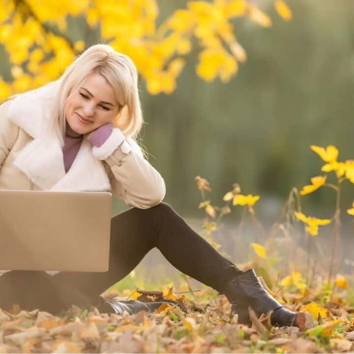 A woman sits on the ground outdoors among autumn leaves, using a laptop and smiling. She is wearing a light coat and boots.