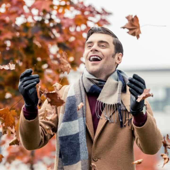 A man wearing a brown coat, scarf, and gloves smiles while tossing autumn leaves in the air outdoors, with orange-leaved trees in the background.