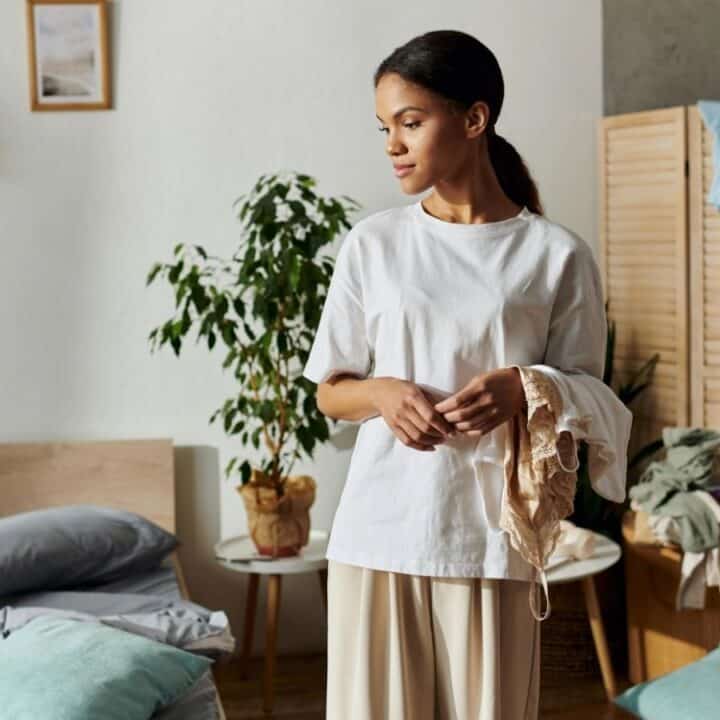A woman stands in a bedroom holding clothes, with a bed, pillows, potted plants, and a folding screen with laundry in the background.
