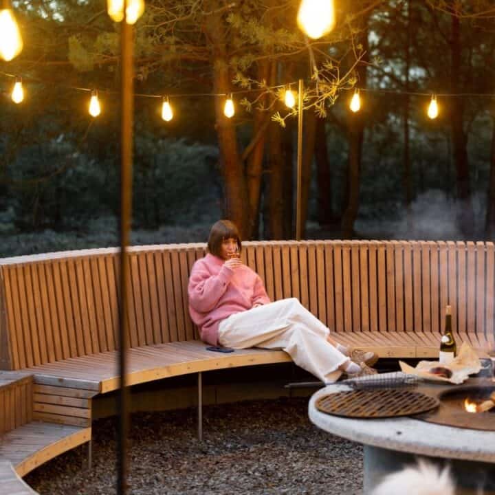 A person in a pink sweater sits on a curved wooden bench by a fire pit, surrounded by string lights in an outdoor wooded area.