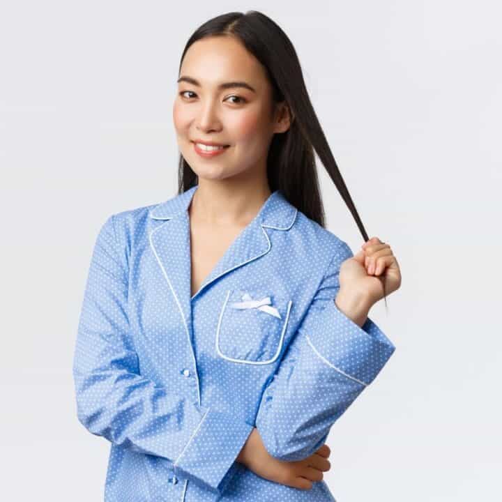 Woman in blue pajamas stands against a plain background, smiling slightly and holding a strand of her long hair with one hand.