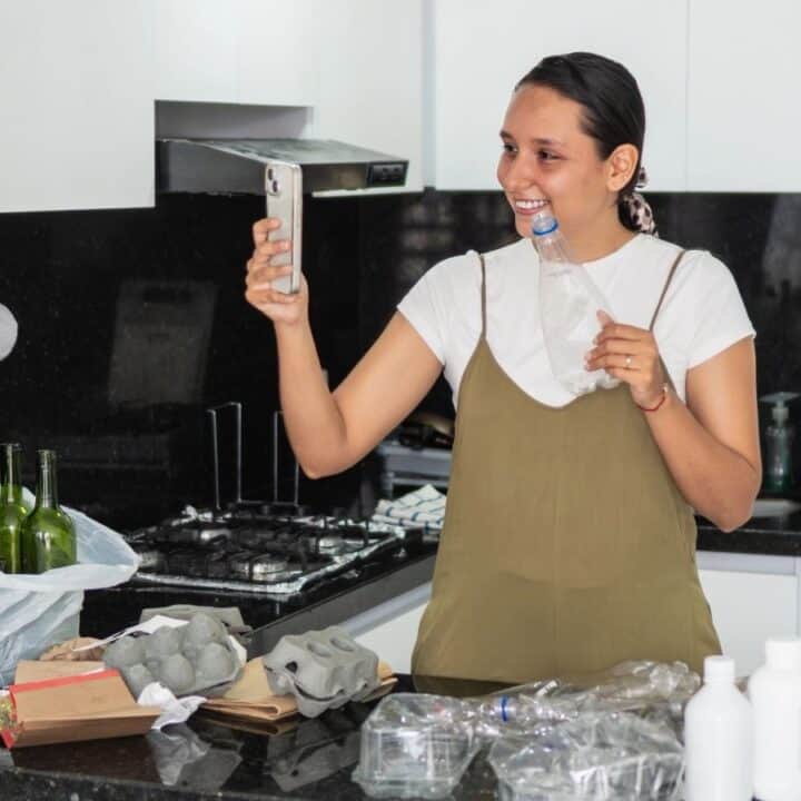 A woman in a kitchen brushes her teeth while holding a phone up for a selfie, surrounded by groceries and kitchen items on the counter.