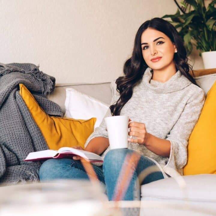 A woman sits on a sofa with yellow cushions, holding a white mug and an open book in her lap, with a gray blanket and a plant in the background.