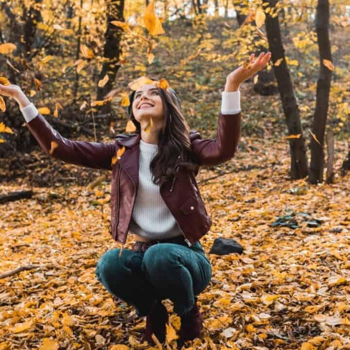 A woman in a maroon jacket crouches on the ground in a forest, smiling and tossing yellow autumn leaves into the air.